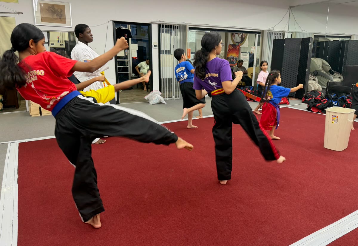 Two girls in black trousers practice kicks across the red mat during class.