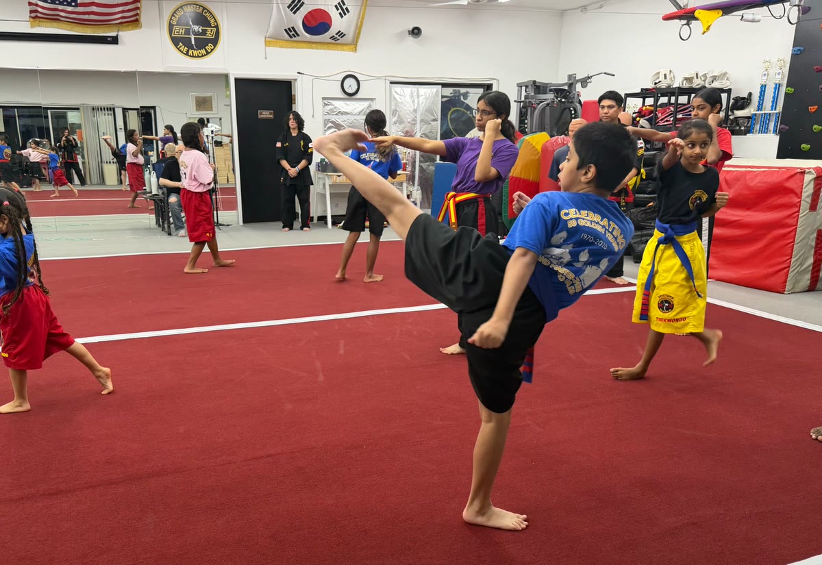 A boy in a blue shirt throws a high kick under the American and Korean flags while classmates practice behind him.