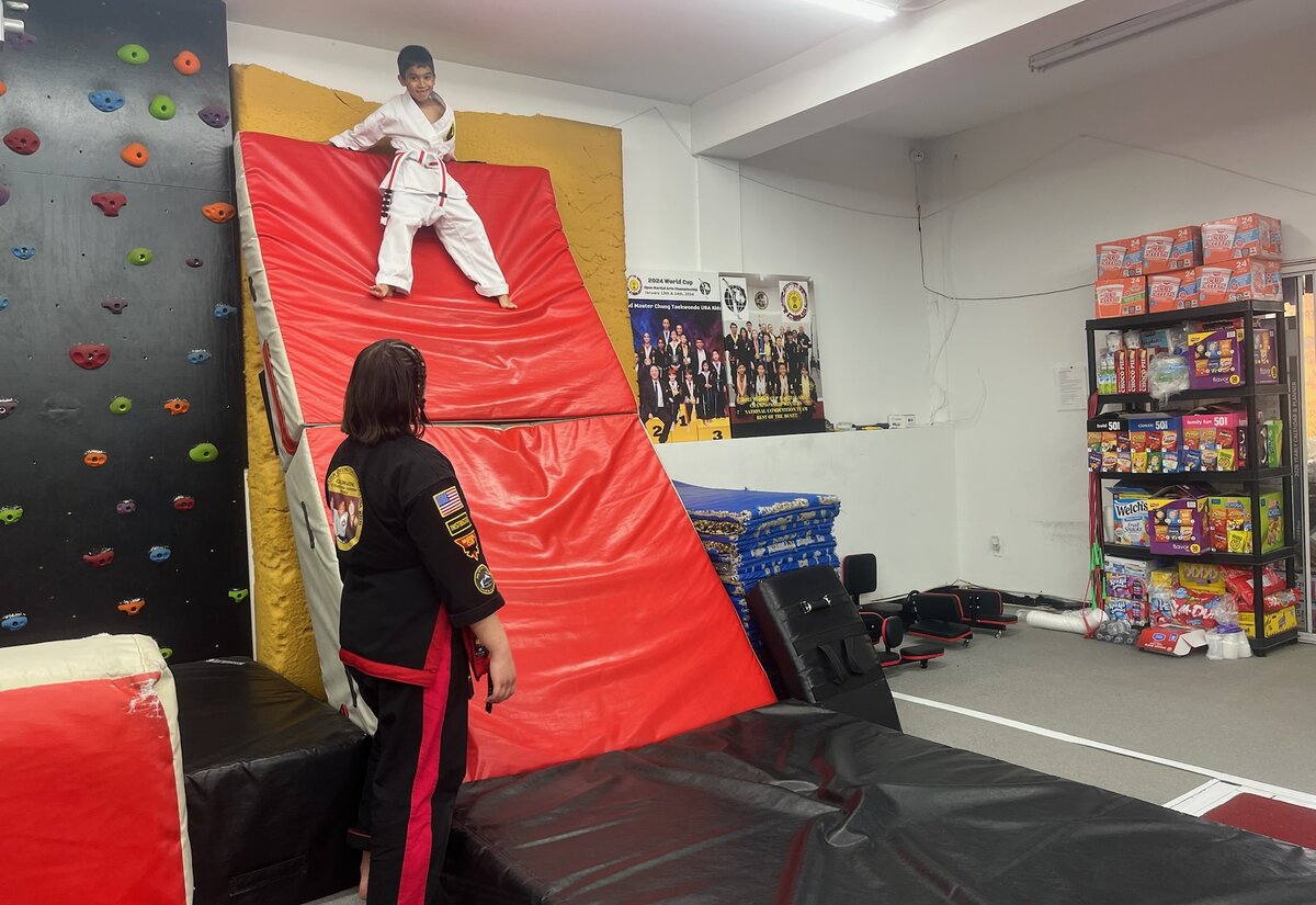 A student in a white uniform stands at the top of a padded climbing ramp while an instructor watches from below.