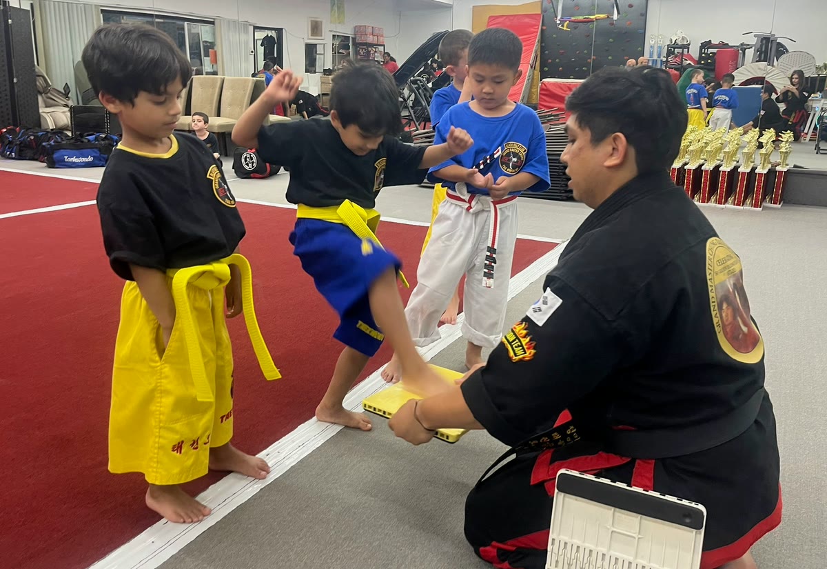 A black belt kneels and holds a breaking board low as a young yellow-belt student stamps down on it, while two classmates wait their turn.