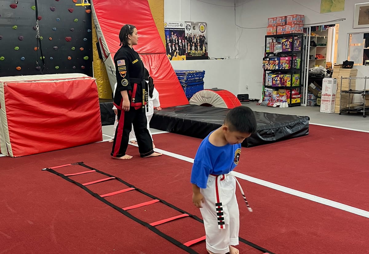 A young student in a blue shirt steps through an agility ladder while an instructor watches.