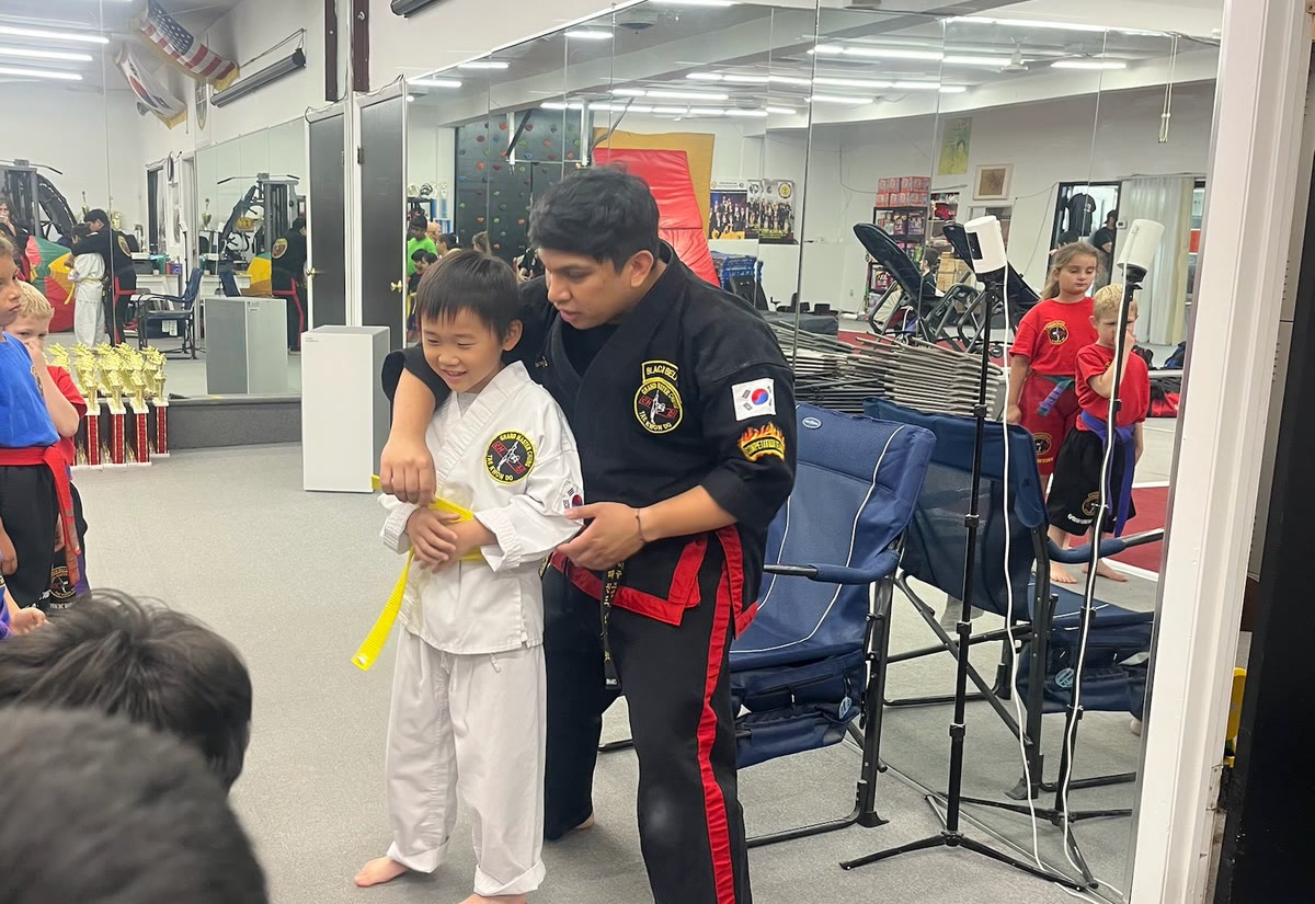 An instructor bends to tie a yellow belt around a young student in a white uniform.