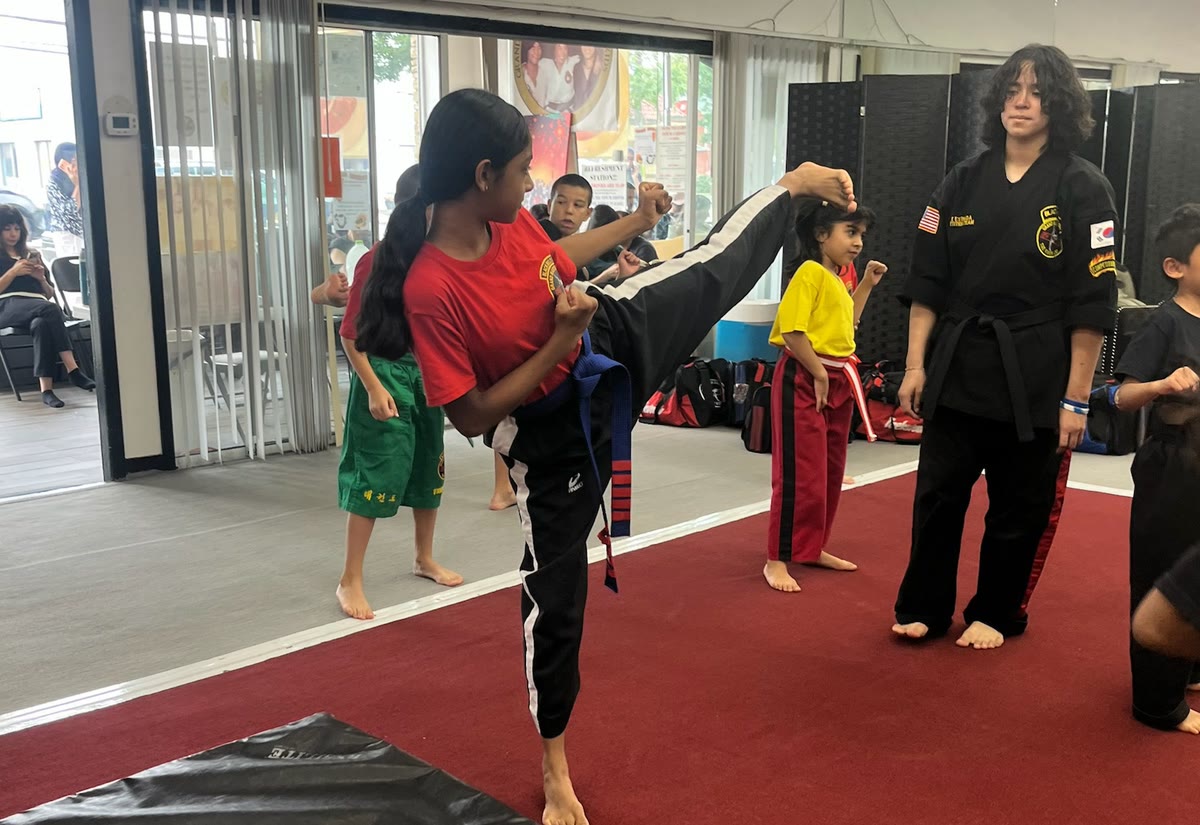 A girl in a red shirt throws a high front kick while an instructor watches.