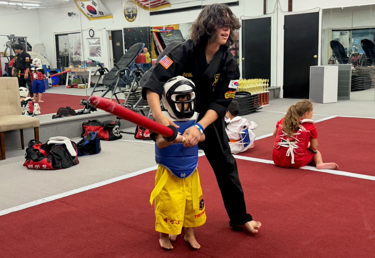 An instructor guides a small child in a helmet and yellow trousers through a foam-stick drill.